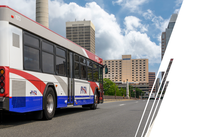 A red, white and blue transit bus on a street headed toward a city.